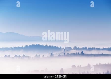 Ciel bleu clair et nuages de brouillard se déplaçant au-dessus du village alpin sur les collines. Paysage tranquille de lever de soleil. Mer de brume dans la vallée. Automne matin ensoleillé dans le mont Banque D'Images