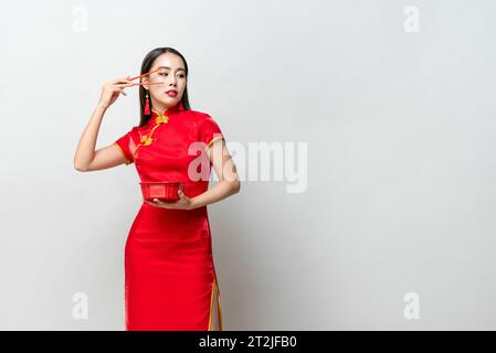 Femme asiatique dans la robe rouge traditionnelle levant le bras avec des baguettes et regardant l'espace de copie de côté sur fond gris Banque D'Images