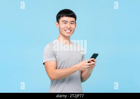 Homme asiatique positif en tenue décontractée texto sur téléphone portable avec sourire en fond bleu clair isolé en studio Banque D'Images