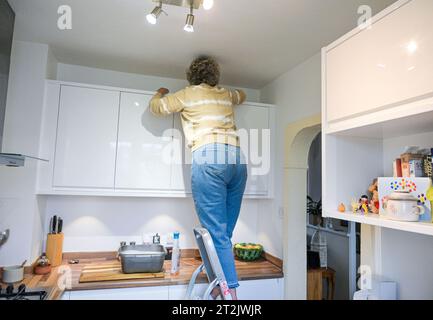 Femme d'âge moyen sur un escabeau nettoyant ses placards de cuisine avec un bol d'eau savonneuse Banque D'Images