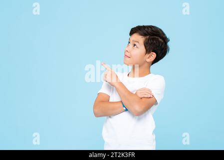 Mignon garçon souriant en t-shirt blanc Uni regardant et pointant la main vers le haut dans le studio isolé fond de couleur bleu clair Banque D'Images