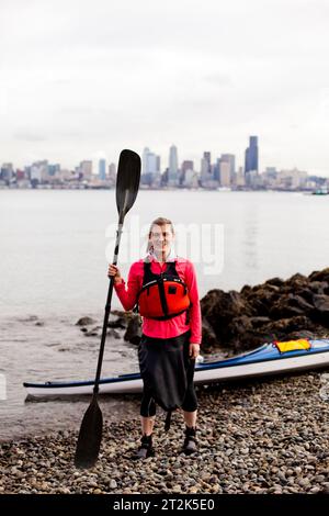 Une jeune fille se tient à côté de son kayak par temps nuageux dans le Puget Sound au large de la côte du centre-ville de Seattle. Banque D'Images