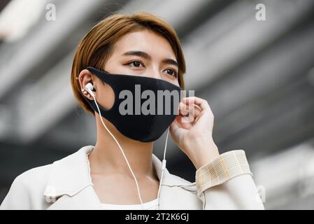 Jeune femme asiatique portant un masque facial à l’extérieur tout en écoutant de la musique dans la ville pendant la pandémie de COVID-19 Banque D'Images