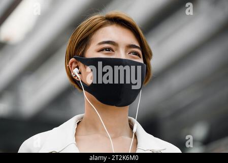 Jeune femme asiatique portant un masque facial à l’extérieur tout en écoutant de la musique dans la ville pendant la pandémie de COVID-19 Banque D'Images