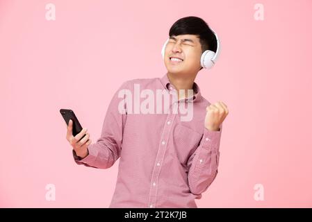 Homme asiatique extatique portant des écouteurs écoutant de la musique en ligne en streaming à partir d'un téléphone portable avec son poing vers le haut dans le fond rose studio Banque D'Images