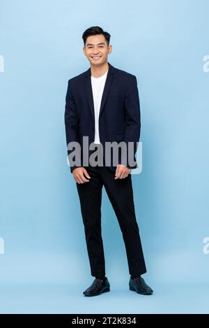 Jeune bel homme asiatique du sud-est portant un costume semi-formel souriant et regardant la caméra dans le fond isolé de studio de couleur bleue Banque D'Images