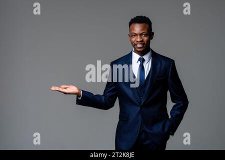 Beau jeune homme d'affaires africain portant costume d'affaires formel souriant et ouvrant la main dans le fond isolé de couleur gris studio Banque D'Images