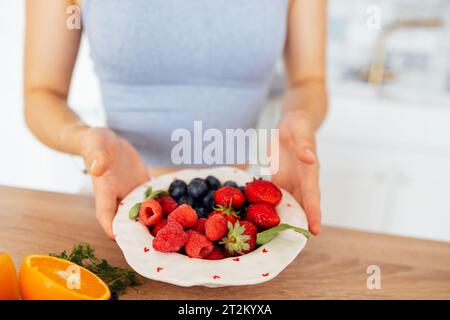 Gros plan des mains féminines tenant un bol en céramique avec des baies saines dans la cuisine. Bleuets, framboises et fraises sur une assiette blanche. Orang Banque D'Images