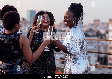 Une fille à la peau foncée verse du champagne dans les verres de ses amis multinationaux. Mignon gens heureux s'amusant à la fête sur le toit. Femmes africaines dans élégant d Banque D'Images