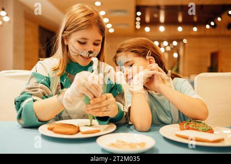Deux filles avec aquagrim sur leurs visages décorent des biscuits de Noël avec un sac à pâtisserie dans un café ou un restaurant. Sœurs en vêtements décontractés ont du plaisir et pa Banque D'Images