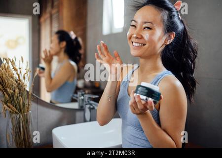 Jeune femme asiatique souriante applique une crème hydratante ou un écran solaire sur son visage tout en se tenant devant un miroir dans la salle de bain à la maison. Charmant gir coréen Banque D'Images