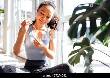 Jeune femme asiatique souriante applique un sérum anti-âge avec des vitamines de la pipette à son visage tout en étant assise dans le salon. Mignonne fille coréenne s'occupe de Banque D'Images