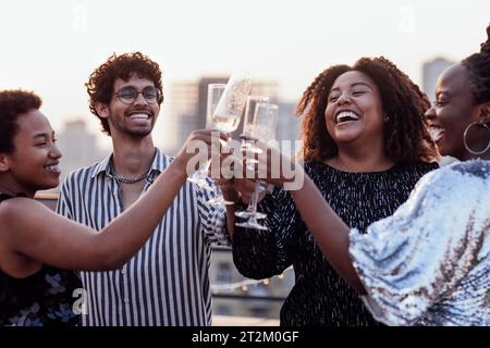 Une fille à la peau foncée verse du champagne dans les verres de ses amis multinationaux. Mignon gens heureux s'amusant à la fête sur le toit. Femmes africaines dans élégant d Banque D'Images