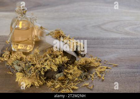 Fleurs séchées de calendula (Calendula officinalis), dans une cuillère en bois sur une tranche de branche d'arbre avec une bouteille d'huile essentielle de calendula. Banque D'Images
