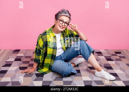 Photo pleine longueur de la femme retraitée détendue satisfaite porter chemise à carreaux assis sur le sol touchant des lunettes isolées sur fond de couleur rose Banque D'Images