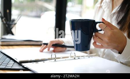 Section médiane de femme d'affaires caucasienne assise au bureau en utilisant un ordinateur portable et tenant une tasse de café. Banque D'Images