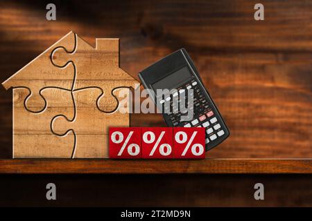 Maison en bois faite de pièces de puzzle, blocs de bois avec des signes de pourcentage rouges et une calculatrice, sur une étagère en bois ou une table avec espace de copie. Banque D'Images