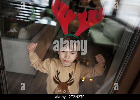 Enfant en chandail tricoté et serre-tête de cerf se tient près de la fenêtre, regarde à l'extérieur. Photo réaliste de style de vie. Banque D'Images