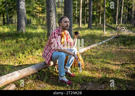 Homme rêveur campeur randonneur assis sur la bûche dans la forêt appréciant boire du thé de thermos ayant la pause. Banque D'Images