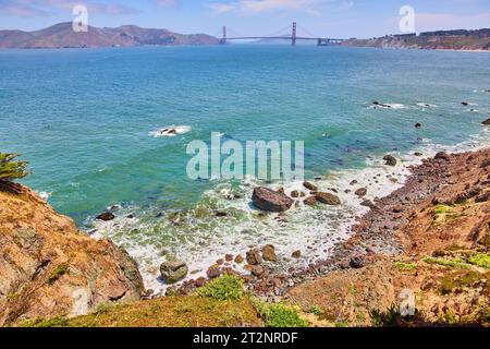 Rivage rocheux avec vagues et mousse de mer tourbillonnant autour des rochers et du lointain Golden Gate Bridge Banque D'Images