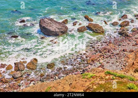 Rivage rocheux avec vagues et mousse de mer tourbillonnant autour des rochers près de colline herbeuse Banque D'Images