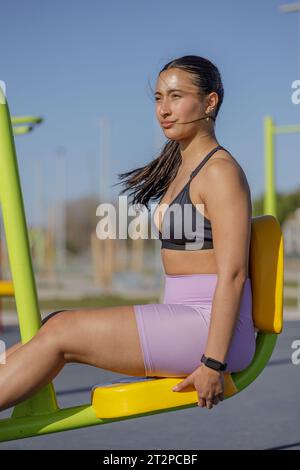 Fille latine exerçant ses jambes sur une machine d'exercice en plein air dans un parc public. Banque D'Images