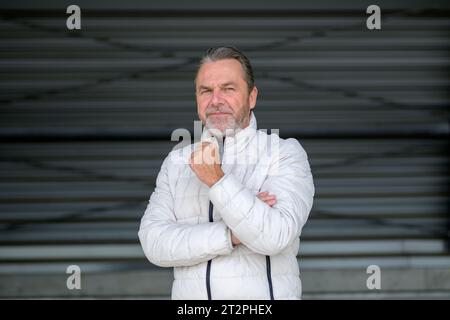 Bel homme aux cheveux gris portant une veste d'hiver blanche avec une main sur sa veste qui semble amicale à la caméra, devant un mur en fer ondulé Banque D'Images