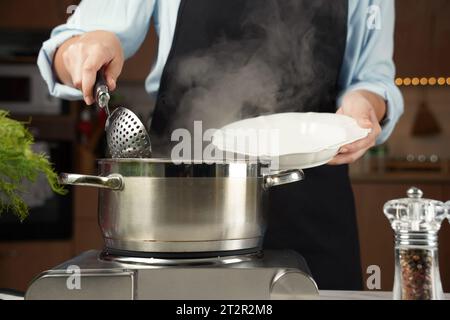Femme avec une louche et une assiette dans les mains. Poêle en acier sur plaque de cuisson électrique. Le processus de cuisson des boulettes Banque D'Images