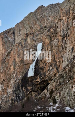 Cascade gelée sur un rocher de falaise haut de couleur brune. Chute de glace effondrée dans les montagnes. Paysage naturel du début du printemps. L'eau a gelé dans la glace. Banque D'Images