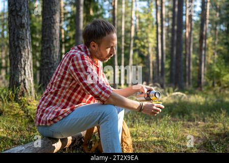 Homme fatigué campeur randonneur assis sur la bûche dans la forêt versant le thé de thermos ayant la pause. Banque D'Images