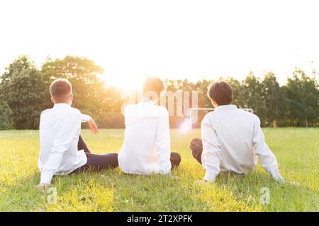 Trois garçons sont assis le dos à nous sur le terrain de football en chemises blanches au coucher du soleil Banque D'Images