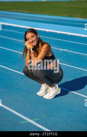 Une belle jeune coureuse, bronzée avec ses longs cheveux bruns, assise sur le côté et accroupie avec ses mains serrées et reposant sa tête sur eux en souriant Banque D'Images