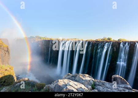 Photo panoramique de la chute d'eau Victoria Falls sur la rivière Zambèze à très haut débit dans la lumière de fin de soirée avec pulvérisation montante et arc-en-ciel intensif au-dessus des chutes Banque D'Images