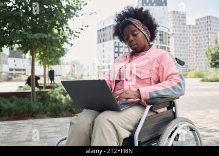 Femme afro-américaine assise en fauteuil roulant dans la ville et utilisant un ordinateur portable Banque D'Images
