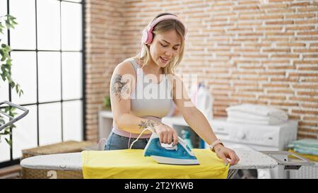 Jeune femme blonde à l'écoute de la musique, repassant des vêtements à la buanderie Banque D'Images