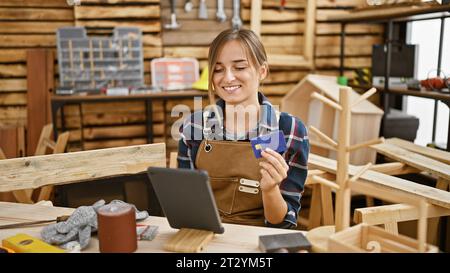 Séduisante jeune femme blonde menuisier souriant tout en payant avec carte de crédit sur touchpad au milieu de l'atelier de menuiserie Banque D'Images