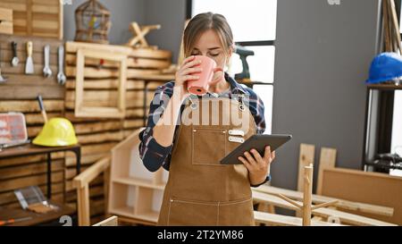 Jolie jeune femme charpentière blonde, café à la main, navigation touchpad au milieu de l'agitation de menuiserie dans l'atelier animé de menuiserie Banque D'Images