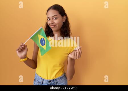 Jeune femme hispanique tenant le drapeau du brésil faisant geste d'argent avec les mains, demandant le paiement du salaire, des affaires millionnaires Banque D'Images