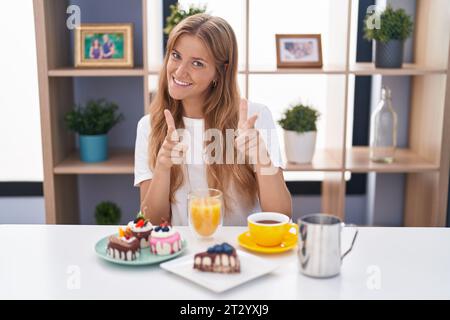 Jeune femme caucasienne mangeant des pâtisseries t pour le petit déjeuner pointant les doigts à la caméra avec le visage heureux et drôle. bonne énergie et vibrations. Banque D'Images