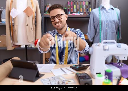 Homme hispanique avec couturier barbe travaillant à l'atelier pointant vers vous et la caméra avec les doigts, souriant positif et joyeux Banque D'Images