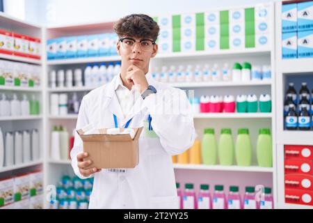 Adolescent hispanique travaillant à la pharmacie pharmacie tenant la boîte avec des pilules visage sérieux pensant à la question avec la main sur le menton, réfléchi à Confus Banque D'Images