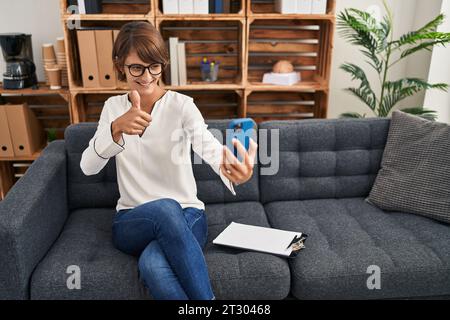 Brunette femme faisant session en ligne au bureau de consultation souriant heureux et positif, pouce vers le haut faisant excellent et signe d'approbation Banque D'Images