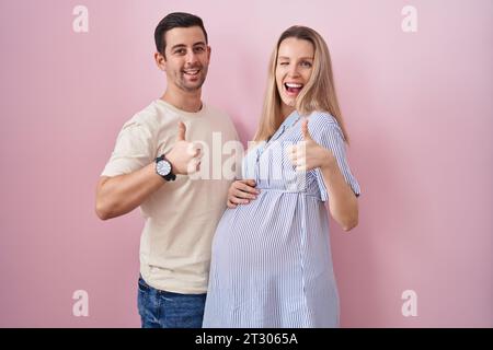 Jeune couple attendant un bébé debout sur fond rose faisant le geste heureux pouces vers le haut avec la main. expression d'approbation regardant l'exposition de la caméra Banque D'Images