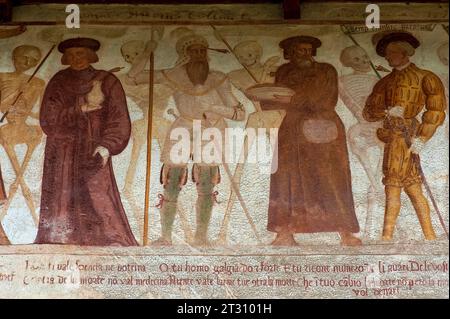 Détail de la frise de fresque Danza Macabra (danse de la mort) du XVIe siècle dans laquelle se dressent des squelettes souriants avec : homme tenant un pot (médecin, apothicaire ou pharmacien), soldat en armure, homme en grand bonnet plat et jeune homme vain ou dandy, chaque mortel percé par une flèche de mort. Le dandy porte un doublet fendu, un tuyau de tronc, des manches bouffées, un collier en dentelle et une plume dans son chapeau. Peint par l'artiste italien Simone II Baschenis (v. 1495-1555) sur le mur extérieur de la Chiesa di San Vigilio à Pinzolo, Trentin-Haut-Adige, Italie. Banque D'Images