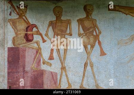 Détail de la frise de fresque Danza Macabra (danse de la mort) du XVIe siècle dans laquelle trio de squelettes se détendent et font de la musique : la mort couronnée (à gauche) joue de la cornemuse et ses compagnons jouent des cornes. Peint par l'artiste italien Simone II Baschenis (v. 1495-1555) sur le mur extérieur de la Chiesa di San Vigilio à Pinzolo, Trentin-Haut-Adige, Italie. Banque D'Images