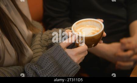 Gros plan d'une femme tenant une tasse de cappuccino et parlant. Film. Jeune couple parle autour d'une tasse de café dans un café. Belle jeune femme tenant Banque D'Images