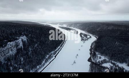 Vue aérienne d'une rivière couverte de neige courbe et d'une vallée boisée. Clip. Rivière courbe gelée coulant parmi une forêt dense. Banque D'Images