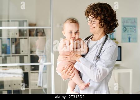 Médecin souriant en manteau blanc tenant bébé sur les bras tout en se tenant au bureau Banque D'Images