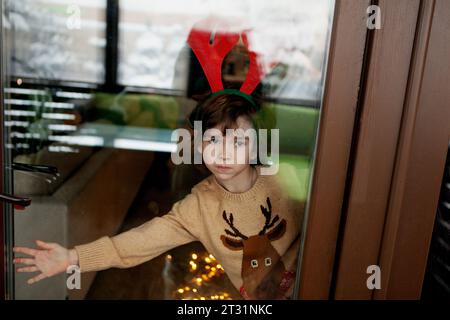 Enfant garçon habillé en costume de renne avec des bois, debout près de la fenêtre, attendant Noël, regardant hors de la maison à travers la vitre de la fenêtre. Banque D'Images