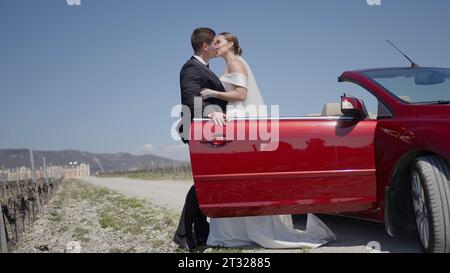 Jeunes mariés près de la voiture cabriolet, sentiments de tendresse. Action. L'homme en costume aide la mariée à quitter la voiture rouge. Banque D'Images